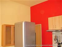 Pohled do kuchyně po renovaci povrchů v béžové a živě červené barvě a světle hnědou kuchyňskou linkou, jen zasunout ledničku